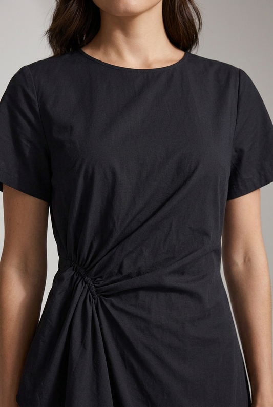 A woman modeling a black midi dress with short sleeves and a twisted waist detail, styled with a straw hat, a woven tote bag, and black strappy sandals against a light gray studio background.