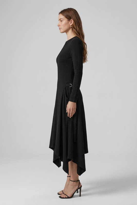 A woman modeling a black long-sleeve midi dress with an asymmetrical high-low hem and side tie details, paired with black strappy heels, against a light gray studio background.