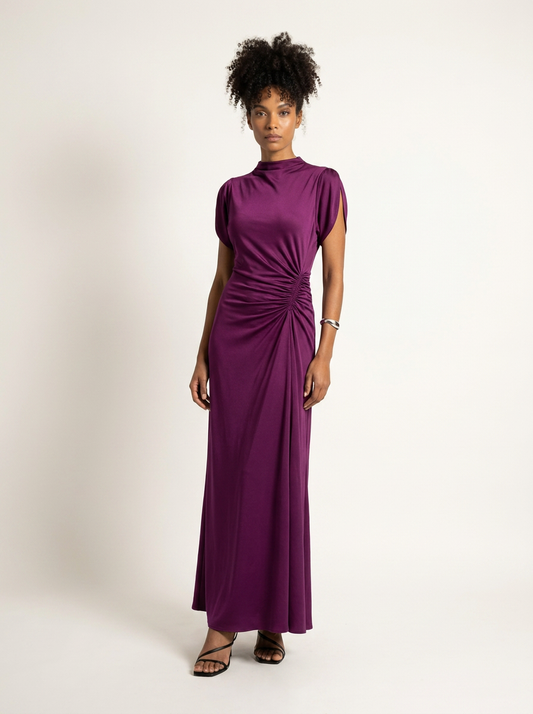 A woman modeling a high-neck, short-sleeved maxi dress in a rich purple shade, standing against a plain off-white studio background.