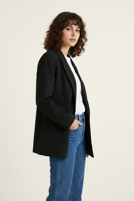 A woman with curly brown hair is shown in profile wearing a black blazer over a white top and blue jeans, standing against a plain light beige studio background.