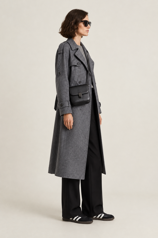 A model wearing a long gray double-breasted coat over a gray top and black wide-leg trousers, accessorized with a black crossbody bag and black sneakers, photographed against a light gray studio background.