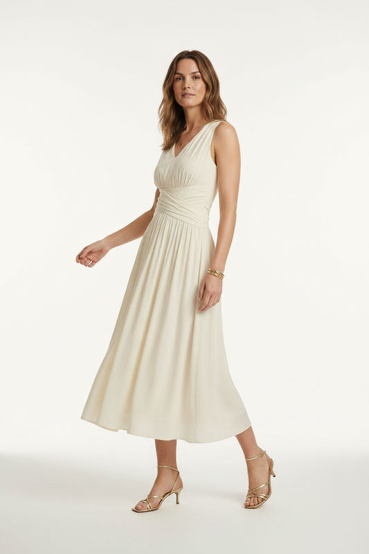 A woman stands on a sunlit Parisian street wearing a sleeveless cream midi dress with a gathered waist and V-neckline, paired with metallic heeled sandals. The background shows classic buildings, a lamppost, and distant street activity.