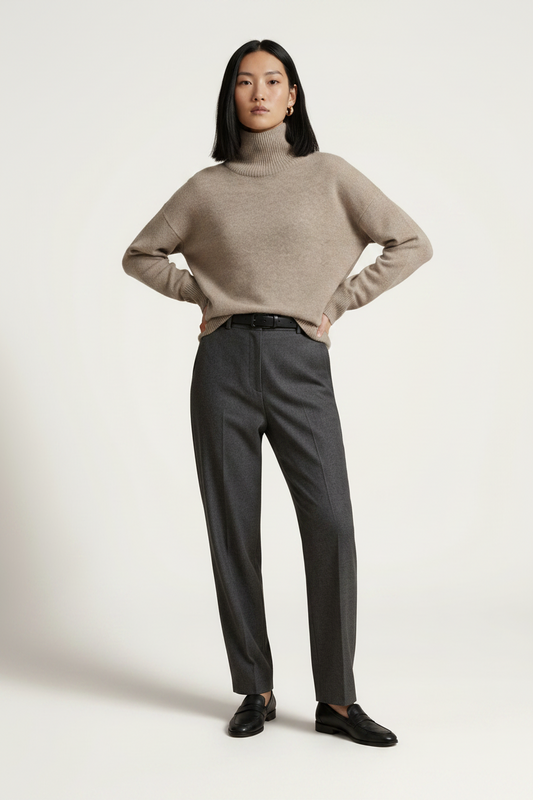 A model stands against a plain light beige studio background wearing a light taupe turtleneck sweater, dark charcoal gray trousers, and black loafers.