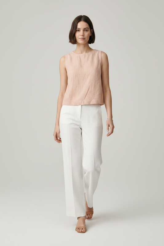 A woman modeling a sleeveless, light blush pink linen top paired with white trousers, shot against a solid gray studio background.