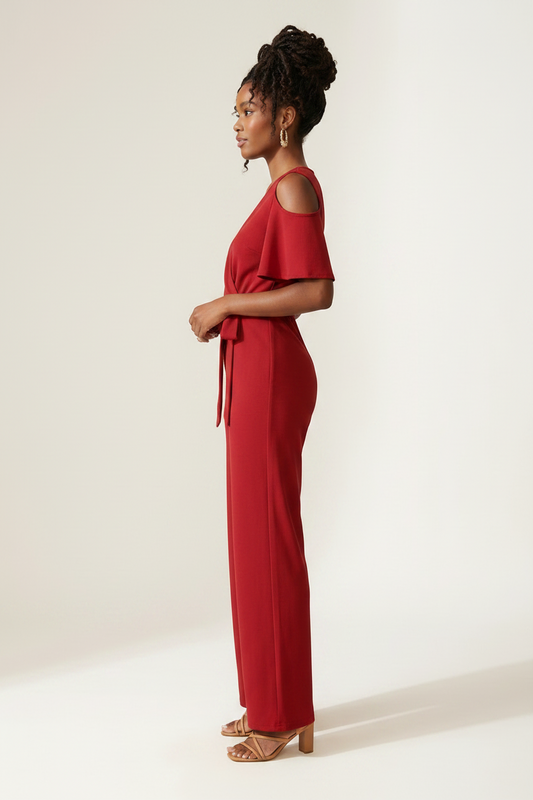 A woman modeling a red cold-shoulder wrap jumpsuit with wide legs and a tie waist, holding a woven straw tote bag against a plain off-white studio background.