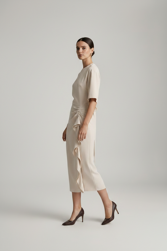 A model stands against a light gray studio background wearing a cream-colored midi dress with short sleeves, a round neckline, and a ruffled side detail, paired with dark brown pointed-toe heels.
