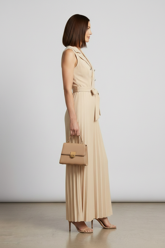 A woman models a sleeveless beige double-breasted jumpsuit with wide legs, pleated detailing, and a matching waist tie. She is holding a matching beige handbag and wearing beige high-heeled sandals against a neutral studio backdrop.