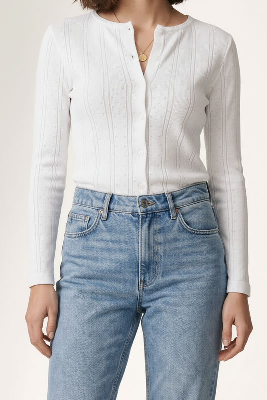 A woman modeling a white ribbed long-sleeve button-front top and light-wash blue jeans with white sneakers, standing against a plain off-white studio background.