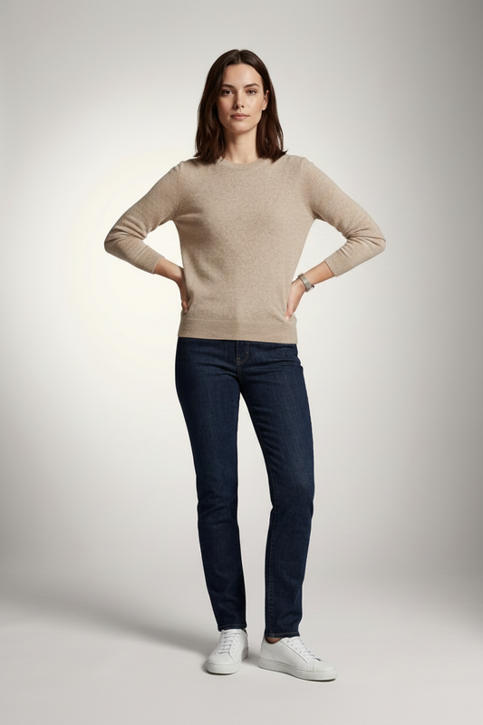 A woman stands against a light gray studio background wearing a beige crewneck sweater, dark blue slim-fit jeans, and white sneakers.