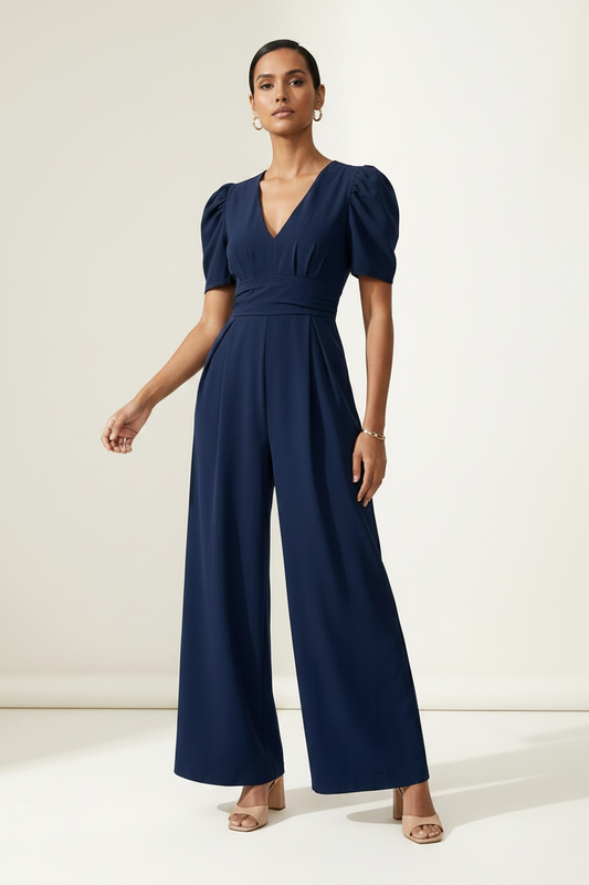 A woman modeling a navy blue jumpsuit with puff sleeves, a wrap-style V-neck bodice, and wide legs, standing against a plain off-white studio background.