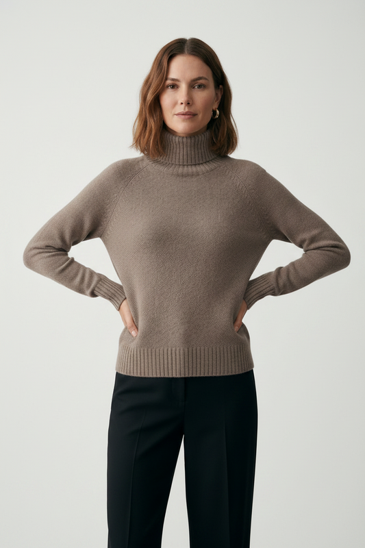 A woman wearing a taupe knit turtleneck sweater and dark trousers, standing against a neutral gray studio background.
