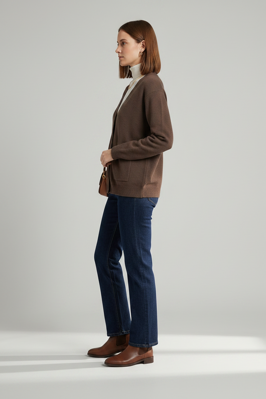 A woman stands against a light gray studio background wearing a brown button-front cardigan over a white turtleneck, dark blue jeans, and brown leather ankle boots, with a small brown shoulder bag.