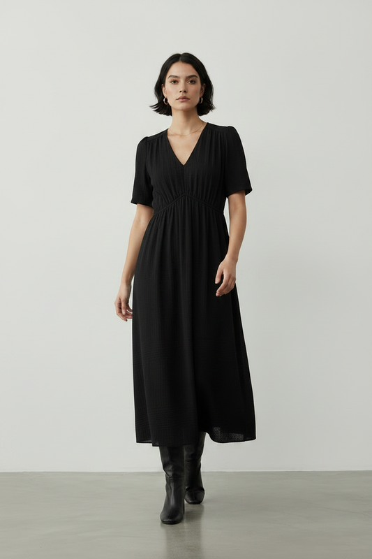 A woman modeling a black midi dress with short sleeves, a V-neckline, and side pockets, paired with black ankle boots, set against a minimalist light gray studio background.