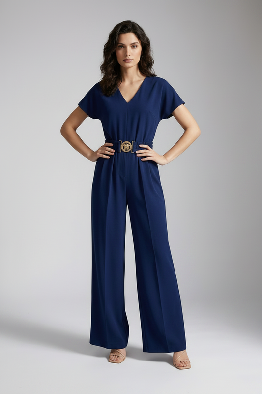 A woman models a navy blue short-sleeve jumpsuit with a gold-tone belt and decorative buckle, styled with a beige clutch and neutral sandals against a light gray studio background.