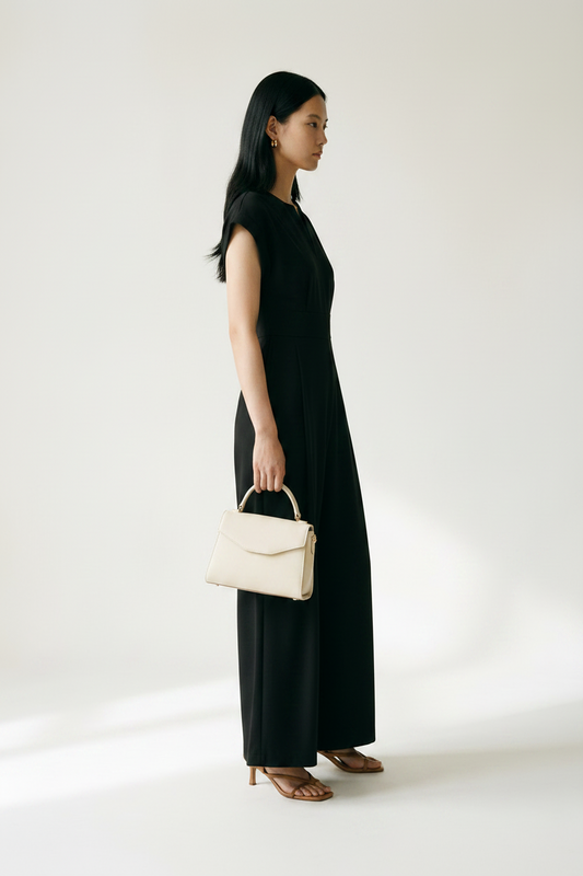 A woman stands in a studio wearing a black sleeveless maxi dress with a split neckline and wide legs, holding a small beige handbag. She is wearing tan heeled sandals against a plain light background.