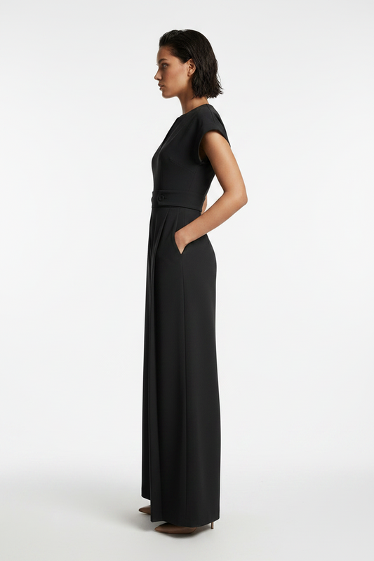 A woman modeling a black wide-leg jumpsuit with short cap sleeves, a split V-neckline, and a matching waist belt, photographed against a neutral gray studio background.