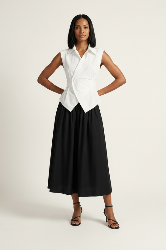 A model stands against a plain light beige studio background wearing a sleeveless white wrap-style shirt and a black midi skirt, holding a small metallic clutch.