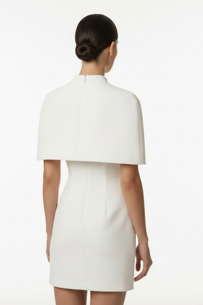 A woman viewed from the back wearing a structured white dress with a high mock neck and a wide cape-like yoke, photographed against a clean white studio background.