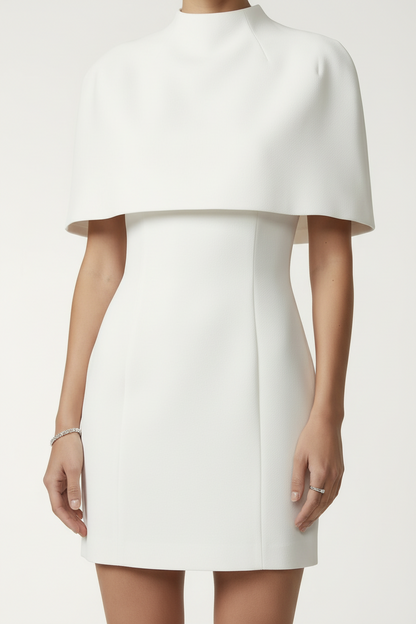 A model stands against a light gray studio background wearing a white high-neck mini dress with a structured cap-like shoulder drape and white pointed-toe heels.