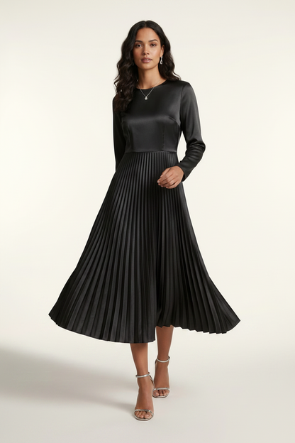 A woman models a black long-sleeve midi dress with a satin-like shiny bodice and a pleated skirt, holding a small silver clutch against a plain light background.