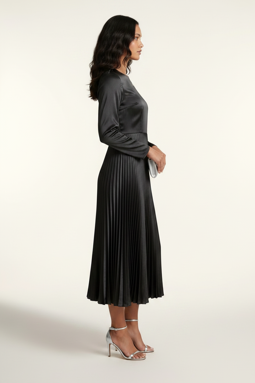 A woman models a black long-sleeve midi dress with a satin-like shiny bodice and a pleated skirt, holding a small silver clutch against a plain light background.