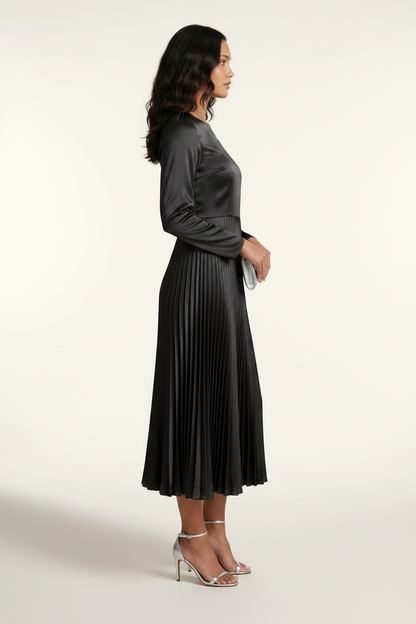 A woman models a black long-sleeve midi dress with a satin-like shiny bodice and a pleated skirt, holding a small silver clutch against a plain light background.