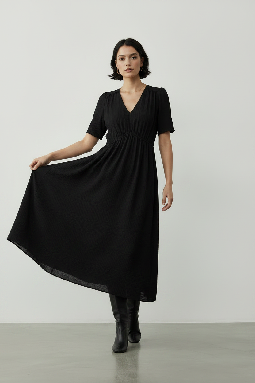 A woman modeling a black midi dress with short sleeves, a V-neckline, and a gathered waist, paired with black ankle boots, set against a minimalist light gray studio background.