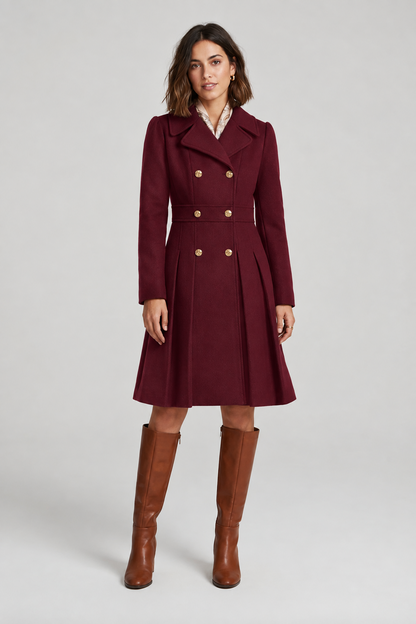 A woman modeling a light pink double-breasted coat with gold buttons, a matching belt, and brown knee-high boots against a light gray studio background.