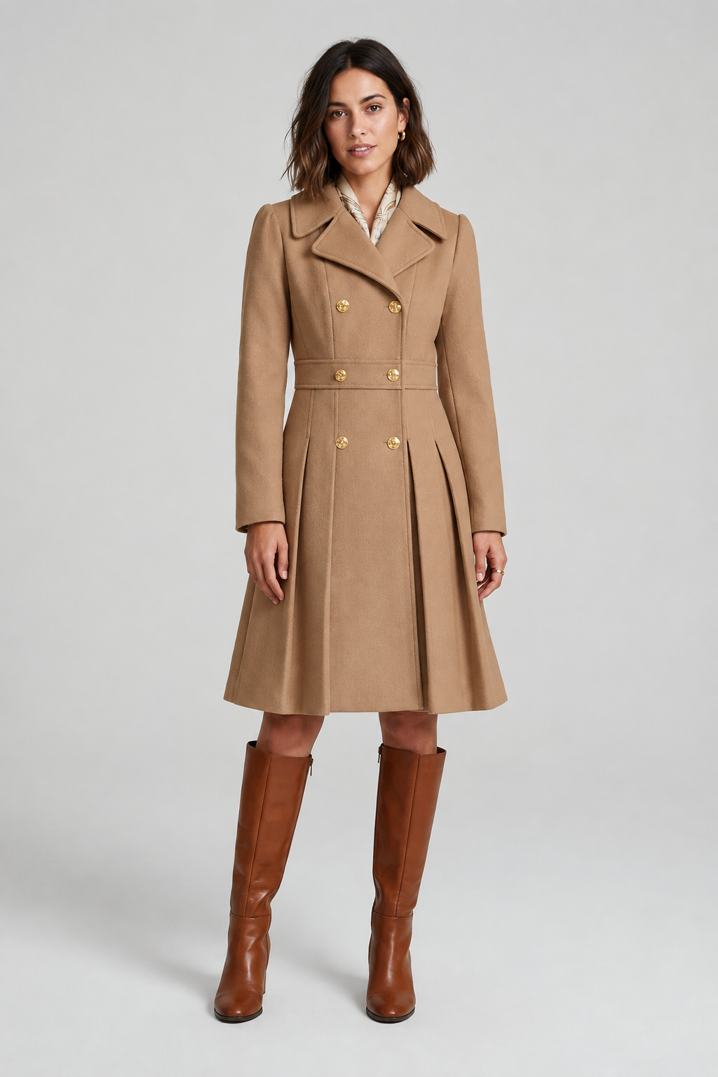 A woman modeling a light pink double-breasted coat with gold buttons, a matching belt, and brown knee-high boots against a light gray studio background.