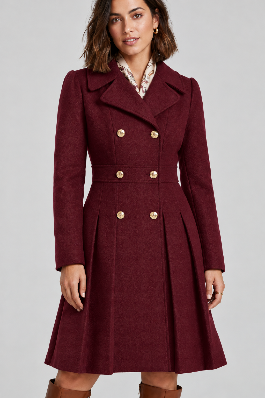 A woman stands against a light gray studio background wearing a knee-length double-breasted coat in deep burgundy with gold-tone buttons, a white collared shirt underneath, and tall brown knee-high boots.