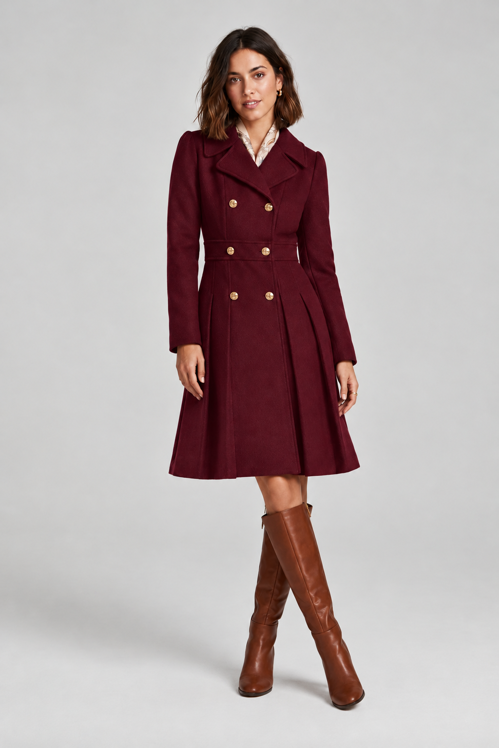 A woman stands against a light gray studio background wearing a knee-length double-breasted coat in deep burgundy with gold-tone buttons, a white collared shirt underneath, and tall brown knee-high boots.
