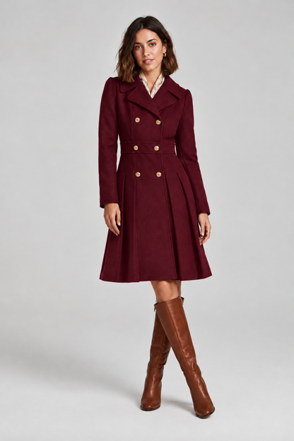 A woman stands against a light gray studio background wearing a knee-length double-breasted coat in deep burgundy with gold-tone buttons, a white collared shirt underneath, and tall brown knee-high boots.
