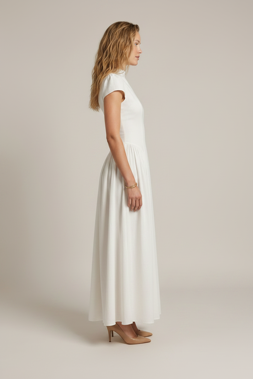 A woman modeling a white high-neck, short-sleeved maxi dress with a gathered waist, standing against a solid light beige studio background.