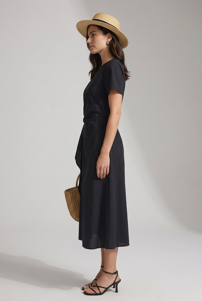 A woman modeling a black midi dress with short sleeves and a twisted waist detail, styled with a straw hat, a woven tote bag, and black strappy sandals against a light gray studio background.