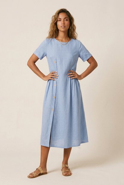 A woman modeling a light blue short-sleeve midi dress with a button-down front, carrying a large woven straw tote bag, against a plain beige studio background.