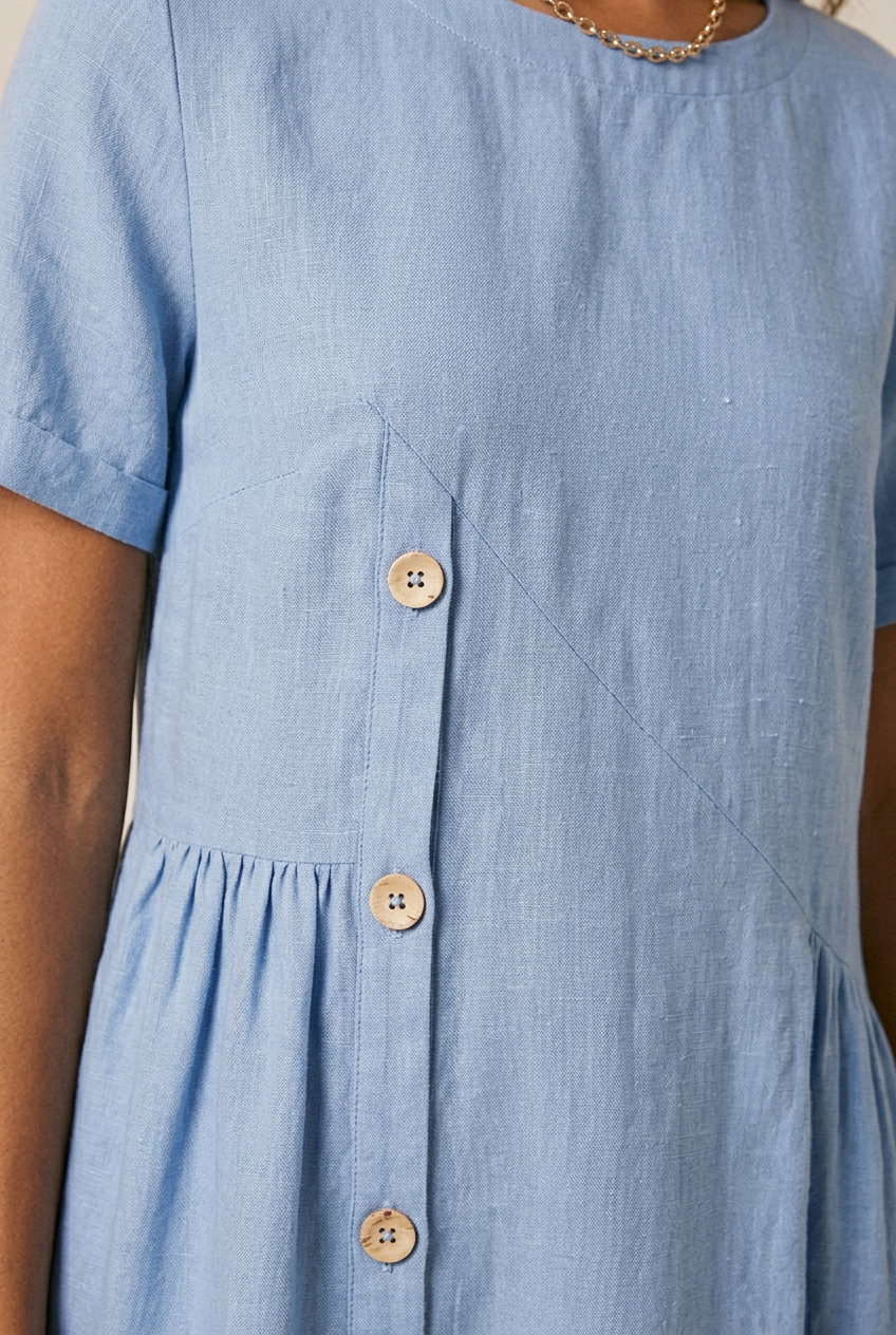 A woman modeling a light blue short-sleeve midi dress with a button-down front, carrying a large woven straw tote bag, against a plain beige studio background.