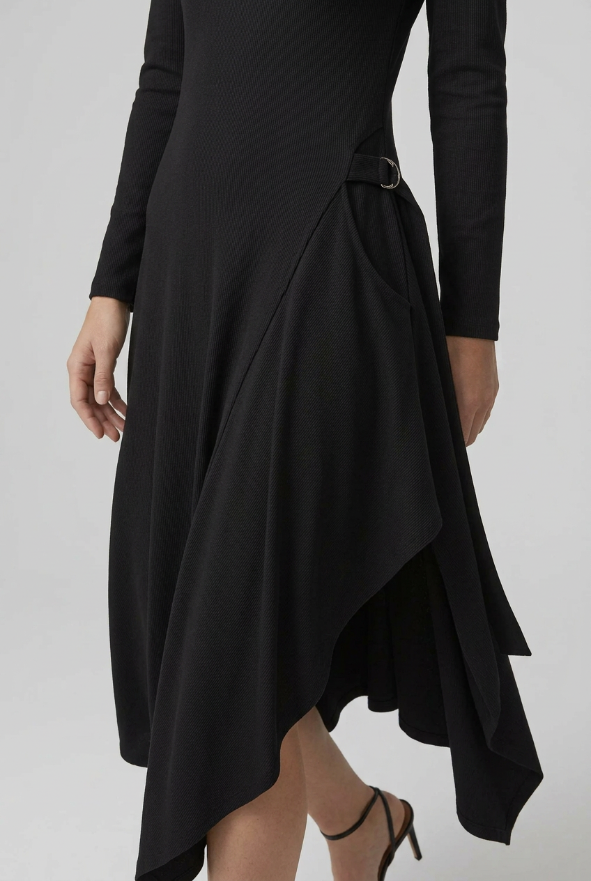 A woman modeling a black long-sleeve midi dress with an asymmetrical high-low hem and a small belt detail at the waist, paired with black strappy heels, against a light gray studio background.