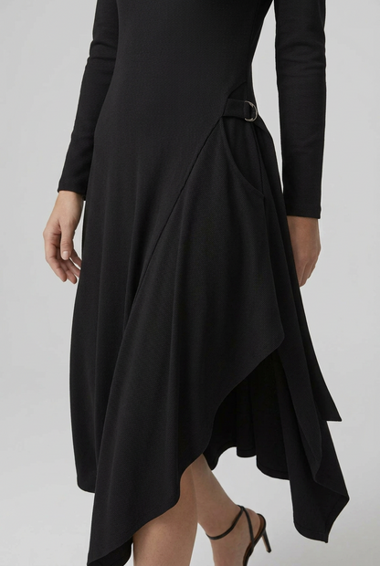 A woman modeling a black long-sleeve midi dress with an asymmetrical high-low hem and a small belt detail at the waist, paired with black strappy heels, against a light gray studio background.