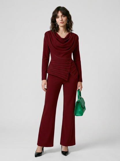 A woman models a matching burgundy two-piece set consisting of a long-sleeve cowl-neck top with a ruched waist and coordinating wide-leg pants, styled with a green handbag and black heels against a light gray studio background.