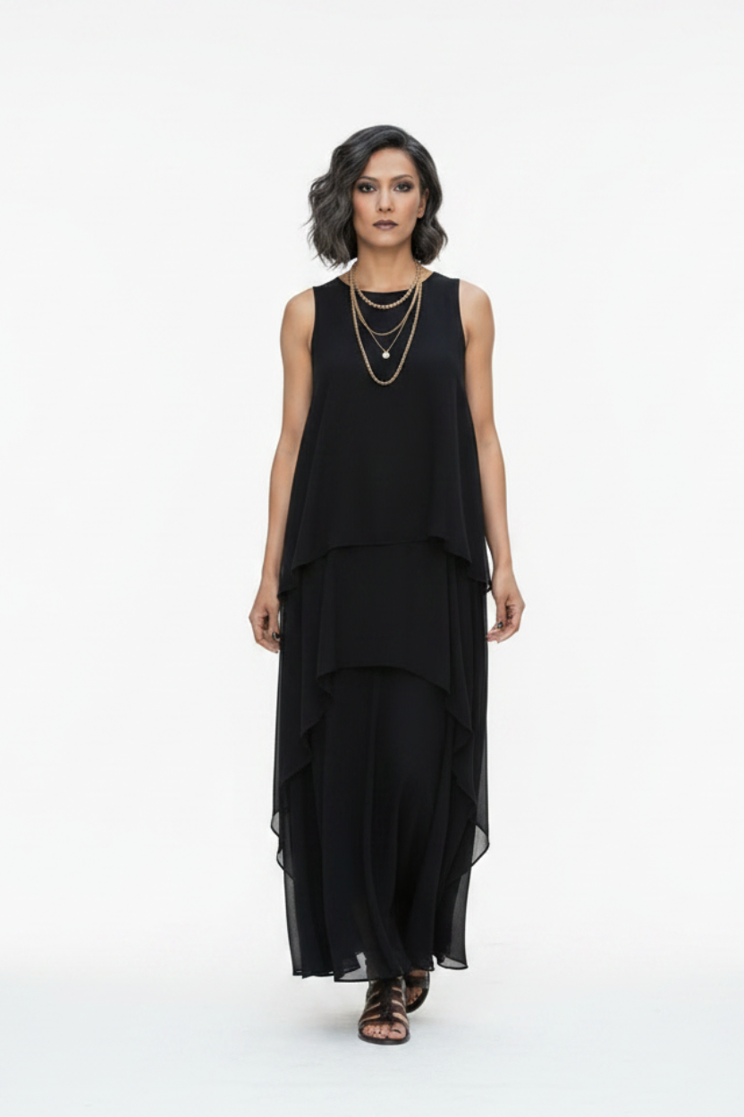 A woman walks down a city street wearing a sleeveless black layered maxi dress with a flowing asymmetrical hem and black strappy sandals.