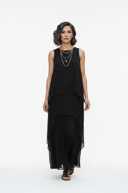 A woman walks down a city street wearing a sleeveless black layered maxi dress with a flowing asymmetrical hem and black strappy sandals.
