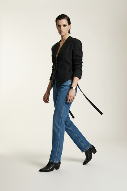 A model stands against a plain light beige studio background wearing a black asymmetrical blazer with side ties, blue jeans, and black ankle boots.