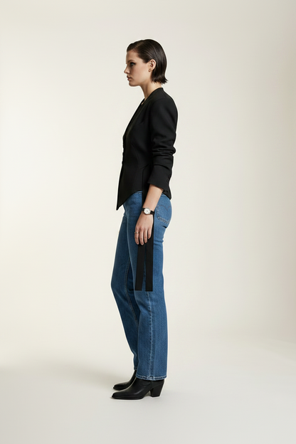 A model stands against a plain light beige studio background wearing a black asymmetrical blazer with side ties, blue jeans, and black ankle boots.
