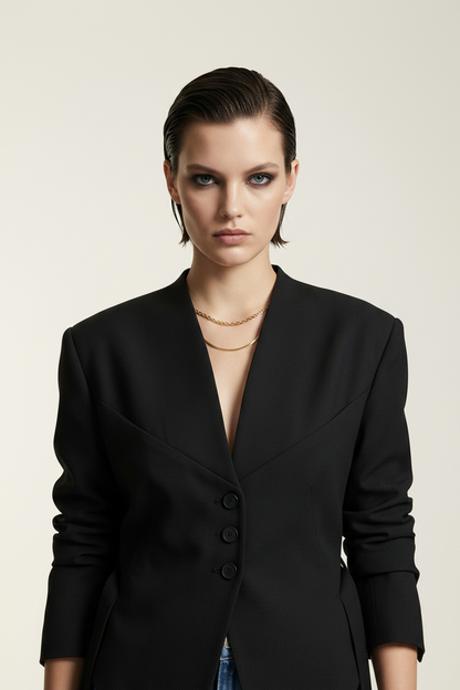 A model stands against a plain light beige studio background wearing a black asymmetrical blazer with side ties, blue jeans, and black ankle boots.
