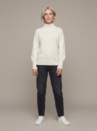 A woman stands against a plain warm gray studio background wearing a cream mock-neck knit sweater, dark charcoal jeans, and white sneakers.