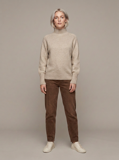 A woman stands against a plain warm gray studio background wearing a light beige knit mock-neck sweater, brown corduroy trousers, and off-white sneakers.