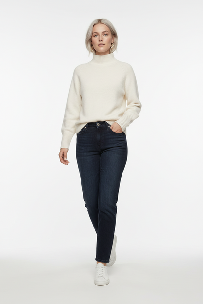 A woman stands against a plain warm gray studio background wearing a cream mock-neck knit sweater, dark charcoal jeans, and white sneakers.