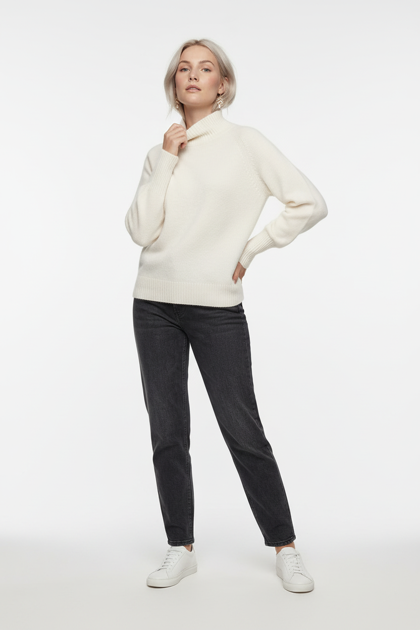 A woman stands against a plain warm gray studio background wearing a cream mock-neck knit sweater, dark charcoal jeans, and white sneakers.