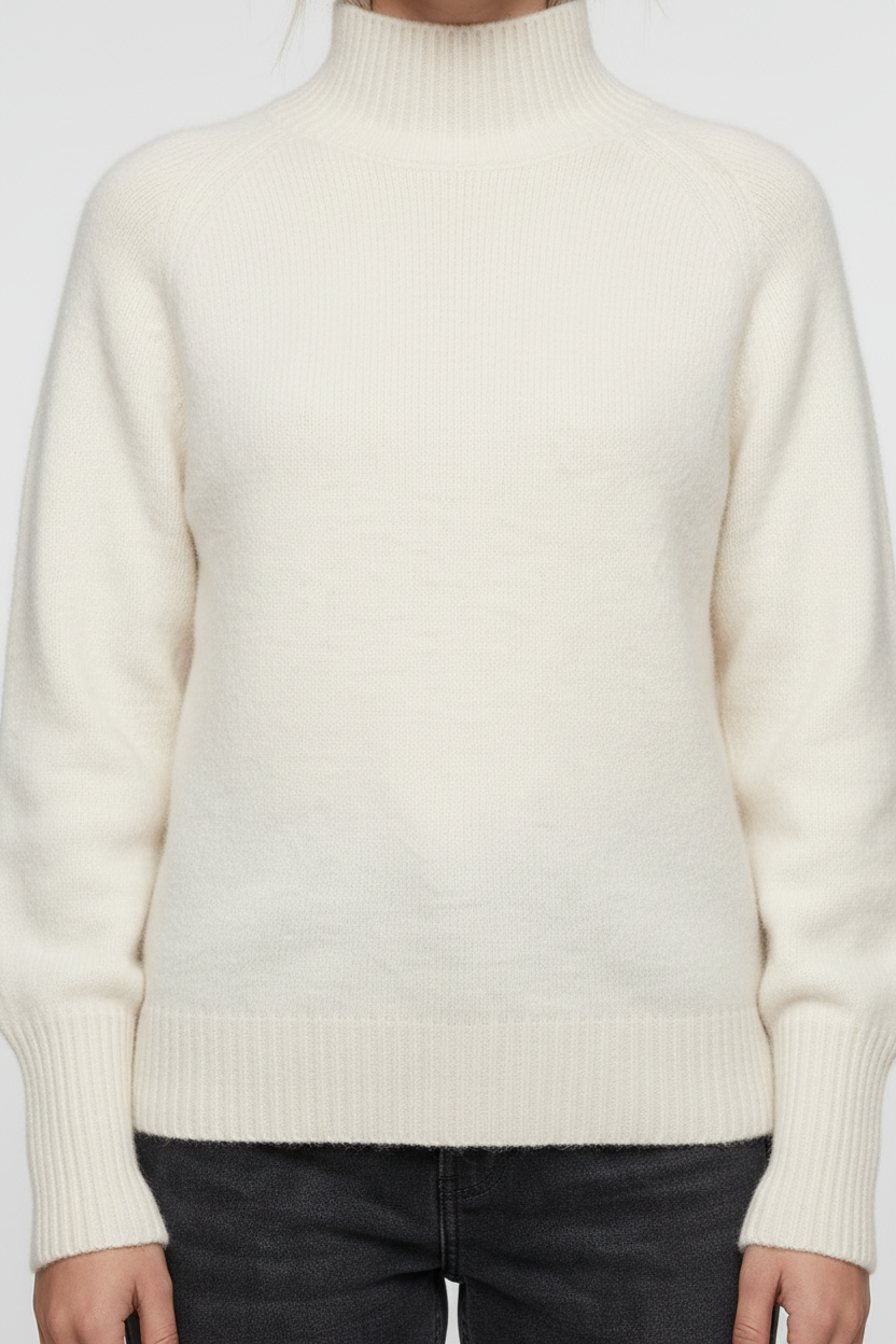 A woman stands against a plain warm gray studio background wearing a cream mock-neck knit sweater, dark charcoal jeans, and white sneakers.