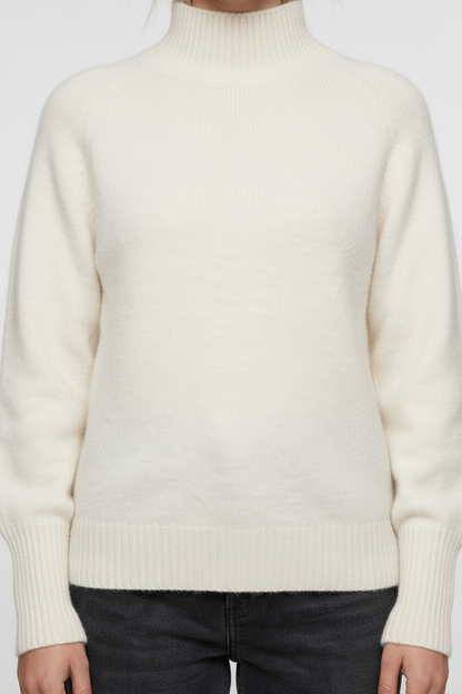 A woman stands against a plain warm gray studio background wearing a cream mock-neck knit sweater, dark charcoal jeans, and white sneakers.